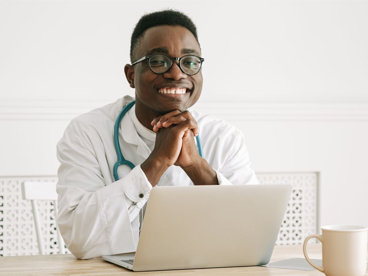 Médico sorridente de jaleco branco com estetoscópio, sentado à mesa com um laptop